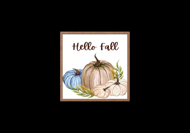 Op de afbeelding: Een ingelijst afdruk met de tekst "Hello Fall" in bruine letters. De afdruk toont een aquarel-illustratie van drie pompoenen: een blauwe, een beige en een witte. De pompoenen worden omringd door groene bladeren en tarwehalmen.