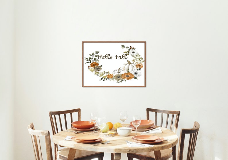 May include: A framed print with a white background and a floral wreath with pumpkins and the text "Hello Fall". The print is hanging on a white wall above a wooden table with four chairs.