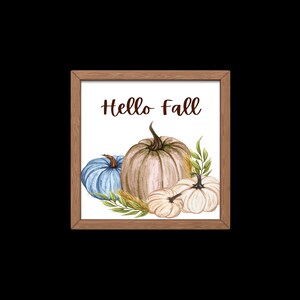 Op de afbeelding: Een ingelijst afdruk met de tekst "Hello Fall" in bruine letters. De afdruk toont een aquarel-illustratie van drie pompoenen: een blauwe, een beige en een witte. De pompoenen worden omringd door groene bladeren en tarwehalmen.