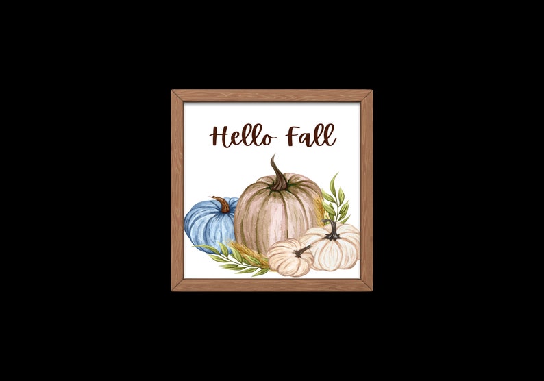 Op de afbeelding: Een ingelijst afdruk met de tekst "Hello Fall" in bruine letters. De afdruk toont een aquarel-illustratie van drie pompoenen: een blauwe, een beige en een witte. De pompoenen worden omringd door groene bladeren en tarwehalmen.