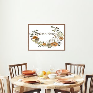 May include: A framed print displaying the words "Autumn Harvest" within a floral wreath of pumpkins and flowers in white, orange, and green. The print is above a round wooden dining table set with plates, bowls, and glasses.