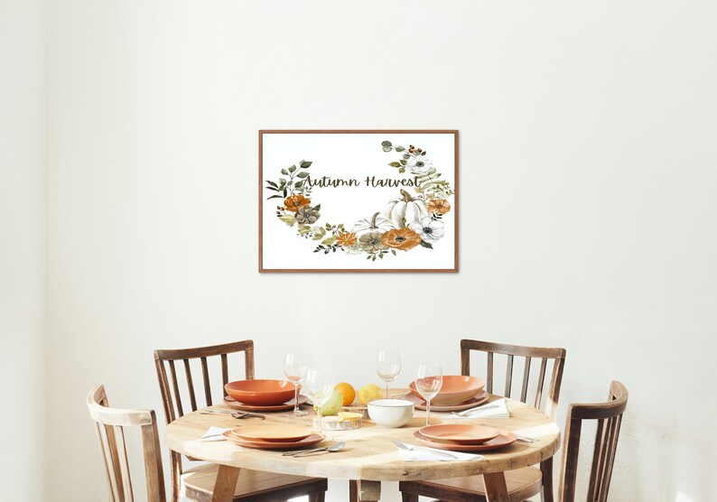 May include: A framed print displaying the words "Autumn Harvest" within a floral wreath of pumpkins and flowers in white, orange, and green. The print is above a round wooden dining table set with plates, bowls, and glasses.