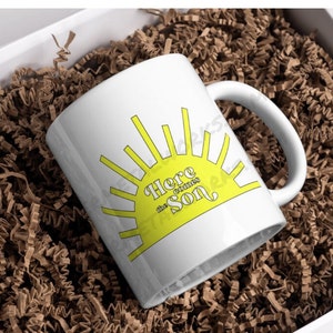 May include: White ceramic mug with a yellow sun graphic and the text "Here comes the sun". The mug is in a white box with brown packing paper.
