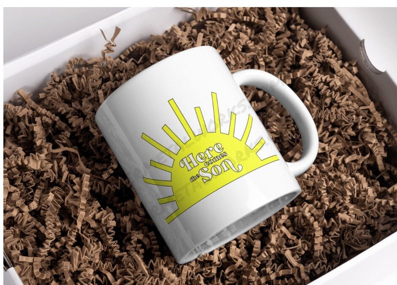 May include: White ceramic mug with a yellow sun graphic and the text "Here comes the sun". The mug is in a white box with brown packing paper.