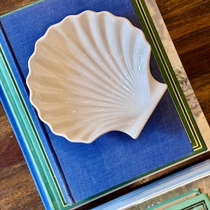 May include: A white ceramic dish shaped like a scallop shell with a glossy finish and radiating ridges. The dish is positioned on top of a blue hardcover book with green and gold trim, which rests above another book with visible text.