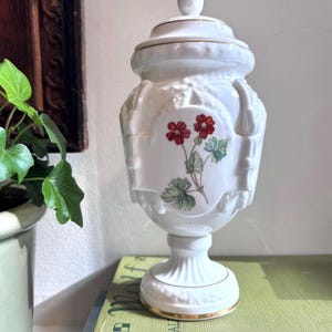 May include: A white porcelain lidded urn featuring a central oval medallion with a botanical illustration of two red flowers and green leaves. The vessel has a pedestal base, embossed relief details, and thin gold trim along the lid and base edges.