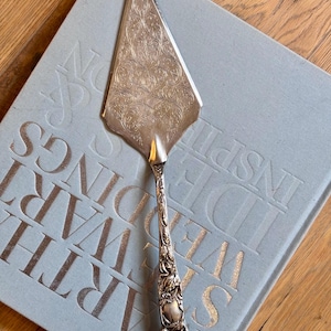 May include: A silver-toned metal cake server featuring an intricate, engraved floral pattern on the triangular blade and a highly detailed, embossed botanical design along the handle, ending in a rounded, ornate finial at the base of the stem.