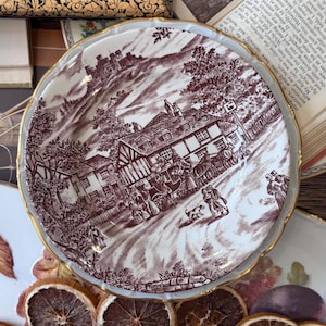 May include: A round ceramic plate with a scalloped edge and a gold-tone rim, featuring a detailed dark brown transferware scene of a horse-drawn carriage in front of a timber-framed building, with a castle on a hill in the background.