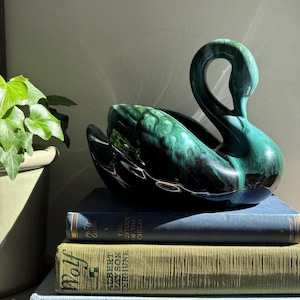 May include: A ceramic swan figurine featuring a glossy finish with blended shades of deep black and vibrant emerald green. The swan is sculpted in a curved, elegant posture with its neck arched forward, resting on a stack of three vintage hardcover books.
