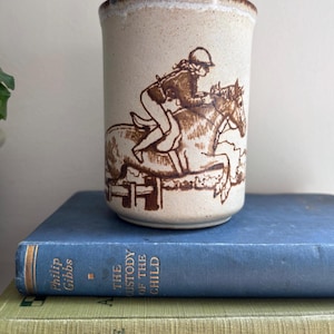 Peut inclure: Un mug cylindrique en céramique avec une finition beige mouchetée et un bord brun. L'avant présente une illustration au trait brun représentant une personne montant un cheval sautant par-dessus une clôture. Le mug a un corps lisse et droit.