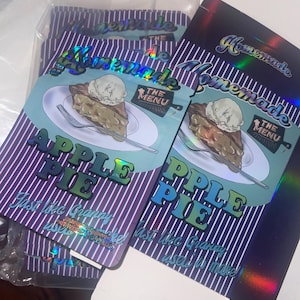 K&ouml;nnte beinhalten: Verpackung f&uuml;r "Homemade Apple Pie" mit einem St&uuml;ck Kuchen und Eis. Das Design enth&auml;lt den Text "Apple Pie" und "Just like Granny used to make!" auf einem Hintergrund aus lila und wei&szlig;en Streifen. Holografische Verpackung.