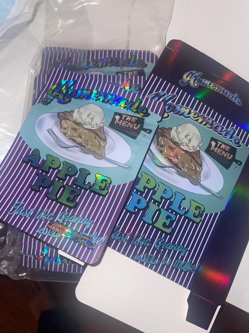 K&ouml;nnte beinhalten: Verpackung f&uuml;r "Homemade Apple Pie" mit einem St&uuml;ck Kuchen und Eis. Das Design enth&auml;lt den Text "Apple Pie" und "Just like Granny used to make!" auf einem Hintergrund aus lila und wei&szlig;en Streifen. Holografische Verpackung.