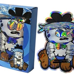 K&ouml;nnte beinhalten: Eine blaue Schachtel und ein holografischer Beutel mit einer Cartoonfigur mit blauem Bandana und braunen F&uuml;&szlig;en. Die Figur hat gro&szlig;e Augen und einen &uuml;berraschten Gesichtsausdruck. Auf der Schachtel steht "High Tolerance" und "PUFFz".