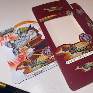 K&ouml;nnte beinhalten: Eine weinrote und wei&szlig;e Produktbox und ein Einsatz mit dem Text "Ken's Tropical Punch" und einer Cartoon-Illustration eines muskul&ouml;sen Charakters. Die Box hat ein Fenster und die Worte "Terps Free!"