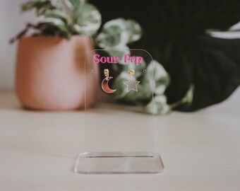 Peut inclure: Présentoir à boucles d'oreilles en acrylique transparent avec le texte "Sour Pop" en rose. Le présentoir contient une paire de boucles d'oreilles, l'une avec un motif de croissant de lune et l'autre avec une étoile. Les boucles d'oreilles ont des accents dorés.