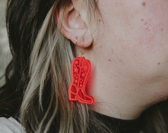 Peut inclure: Boucles d'oreilles en forme de botte de cowboy rouge vif avec des crochets dorés. Les boucles d'oreilles sont de couleur rouge uni avec des découpes en forme d'étoiles. Un accessoire amusant pour les amateurs d'esthétique western.
