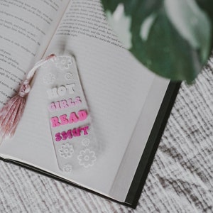 Peut inclure: Un marque-page en acrylique transparent avec le texte "HOT GIRLS READ SMUT" en lettres roses et blanches, orn&eacute; de motifs floraux blancs. Le marque-page a un pompon rose et repose sur les pages d'un livre ouvert.