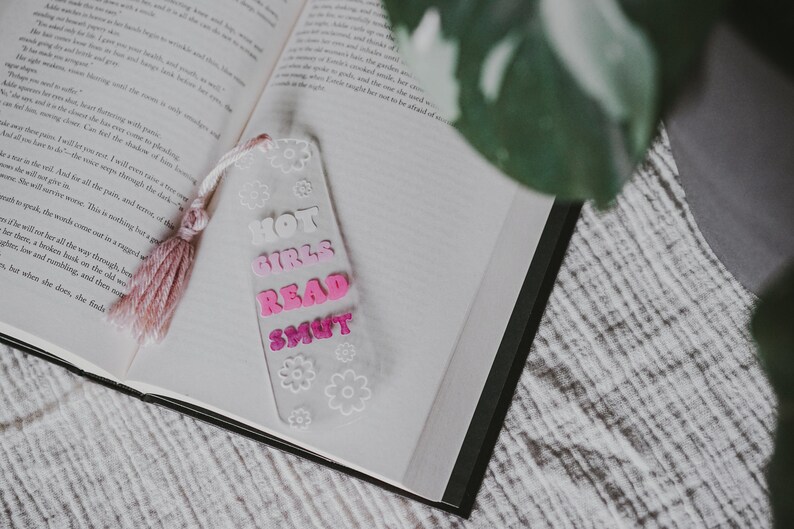 Peut inclure: Un marque-page en acrylique transparent avec le texte "HOT GIRLS READ SMUT" en lettres roses et blanches, orn&eacute; de motifs floraux blancs. Le marque-page a un pompon rose et repose sur les pages d'un livre ouvert.