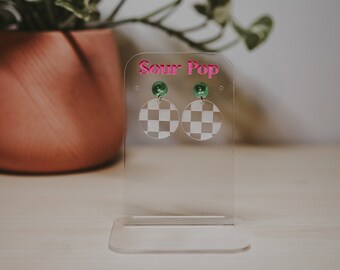 Peut inclure: Présentoir à boucles d'oreilles en acrylique transparent avec le texte "Sour Pop" en rose. Le présentoir contient deux boucles d'oreilles à motif de damier avec des accents verts. Un pot rose avec une plante est en arrière-plan.