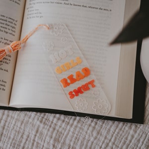 Peut inclure: Marque-page en acrylique transparent avec l'inscription "HOT GIRLS READ SMUT" en lettres orange et jaune. Le marque-page est orn&eacute; d'un pompon et de motifs floraux. Il est plac&eacute; dans un livre ouvert.