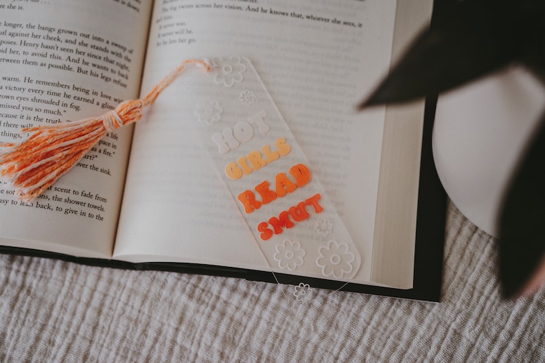 Peut inclure: Marque-page en acrylique transparent avec l'inscription "HOT GIRLS READ SMUT" en lettres orange et jaune. Le marque-page est orn&eacute; d'un pompon et de motifs floraux. Il est plac&eacute; dans un livre ouvert.