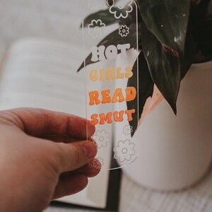 Peut inclure: Marque-page en acrylique transparent avec le texte "HOT GIRLS READ SMUT" en orange et jaune r&eacute;tro. Le marque-page a un motif floral et est tenu devant une plante.
