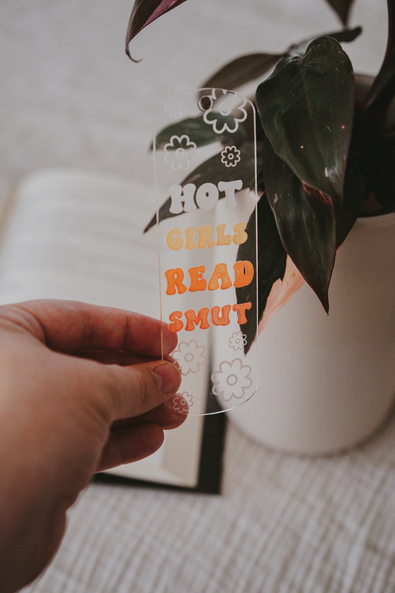 Peut inclure: Marque-page en acrylique transparent avec le texte "HOT GIRLS READ SMUT" en orange et jaune r&eacute;tro. Le marque-page a un motif floral et est tenu devant une plante.