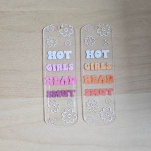Peut inclure: Deux marque-pages en acrylique transparent avec les mots "HOT GIRLS READ SMUT" dans des lettres color&eacute;es de style r&eacute;tro. Des motifs de fleurs blanches sont dispers&eacute;s sur les marque-pages. Les marque-pages sont sur une surface en bois clair.