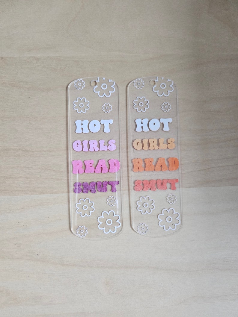 Peut inclure: Deux marque-pages en acrylique transparent avec les mots "HOT GIRLS READ SMUT" dans des lettres color&eacute;es de style r&eacute;tro. Des motifs de fleurs blanches sont dispers&eacute;s sur les marque-pages. Les marque-pages sont sur une surface en bois clair.