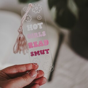 Peut inclure: Marque-page en acrylique transparent avec un pompon rose. Le marque-page pr&eacute;sente le texte "HOT GIRLS READ SMUT" dans une police r&eacute;tro, avec des graphiques de fleurs. Le marque-page est tenu dans une main.