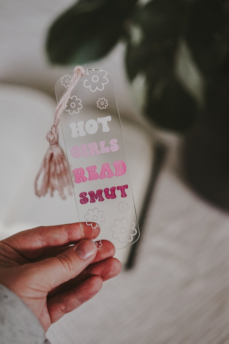 Peut inclure: Marque-page en acrylique transparent avec un pompon rose. Le marque-page pr&eacute;sente le texte "HOT GIRLS READ SMUT" dans une police r&eacute;tro, avec des graphiques de fleurs. Le marque-page est tenu dans une main.