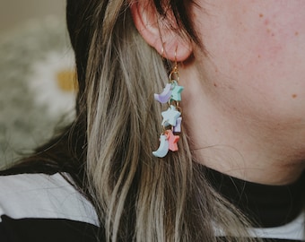 Peut inclure: Boucles d'oreilles pendantes sur le thème céleste. Les boucles d'oreilles sont dotées de crochets dorés et sont ornées de breloques en forme d'étoiles et de croissants de lune aux couleurs pastel. Les breloques sont dans les tons de bleu, de violet et de rose.