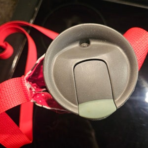 Pu&ograve; includere: Primo piano di un coperchio di una tazza da viaggio grigia con un cinturino rosso. Il coperchio ha una piccola apertura rotonda e uno slider per aprire e chiudere l'apertura.