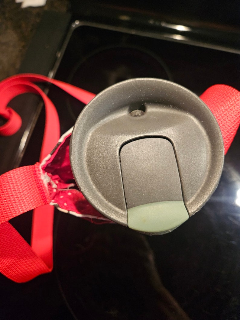 Pu&ograve; includere: Primo piano di un coperchio di una tazza da viaggio grigia con un cinturino rosso. Il coperchio ha una piccola apertura rotonda e uno slider per aprire e chiudere l'apertura.