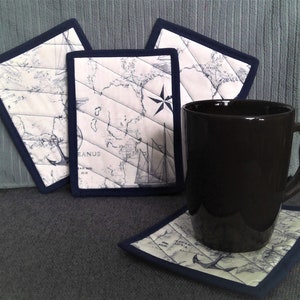 May include: Four blue and white nautical themed fabric coasters with a black coffee mug on top of one of the coasters. The coasters feature a map design with an anchor and a sailboat.
