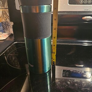 Pu&ograve; includere: Una tazza da viaggio verde e nera con un coperchio nero e un manico nero. La tazza &egrave; alta 17,8 cm e si trova su una superficie nera. La tazza ha la scritta "ntigo" stampata sul lato.