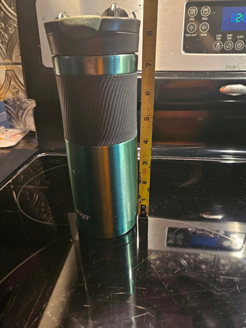 Pu&ograve; includere: Una tazza da viaggio verde e nera con un coperchio nero e un manico nero. La tazza &egrave; alta 17,8 cm e si trova su una superficie nera. La tazza ha la scritta "ntigo" stampata sul lato.