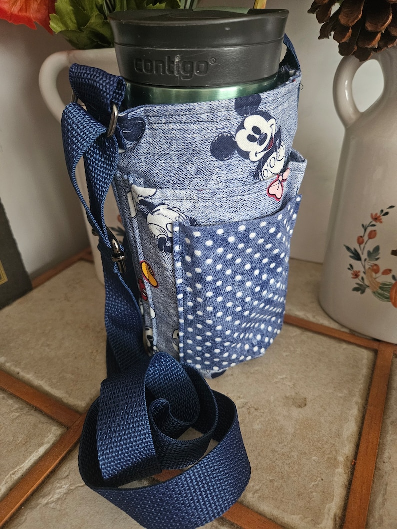 Pu&ograve; includere: Un porta-bottiglia in tessuto blu e bianco con un design di Topolino, una bottiglia d'acqua del marchio Contigo e una tracolla regolabile blu scuro.