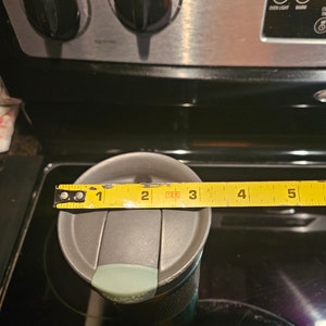 Pu&ograve; includere: Una tazza da viaggio grigia con un coperchio verde acqua e un corpo con motivo nero e grigio. La tazza viene misurata con un metro a nastro giallo, che mostra una misura di 3 pollici.