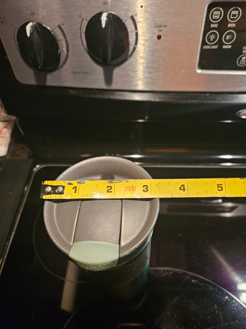 Pu&ograve; includere: Una tazza da viaggio grigia con un coperchio verde acqua e un corpo con motivo nero e grigio. La tazza viene misurata con un metro a nastro giallo, che mostra una misura di 3 pollici.