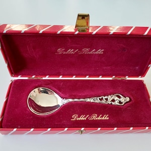 May include: A silver spoon with an ornate handle is displayed in a red and white striped box with a red velvet interior. The box is labelled "Dobbel Rokokko".