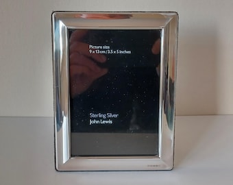 May include: A rectangular sterling silver picture frame with a polished finish. The frame has a black interior and text that reads "Picture size 9 x 13cm / 3.5 x 5 inches" and "Sterling Silver John Lewis".