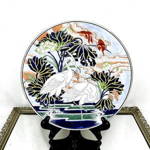 Puede incluir: Un plato de cerámica blanco con un borde azul y dorado. El plato presenta un diseño pintado a mano de dos grullas blancas de pie en un estanque con flores y hojas de loto. El fondo es azul claro con nubes blancas.