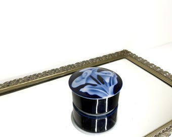 Peut inclure: Petite boîte à bijoux ronde en céramique bleu foncé avec couvercle. Le couvercle présente un motif floral dans les tons de bleu et blanc. La boîte est posée sur une surface miroir dans un cadre doré décoratif.
