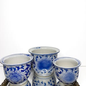 Puede incluir: Tres maceteros de cerámica con base blanca y motivos florales azules. Los maceteros tienen una forma redondeada con un borde acampanado y están dispuestos en grupo, siendo el macetero central el más alto de los tres recipientes a juego.