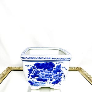 Puede incluir: Una maceta cuadrada de cerámica blanca con un diseño de uvas azules. La maceta tiene un borde azul decorativo y está sobre una superficie reflectante. La maceta mide aproximadamente 10 cm de ancho y 10 cm de largo.