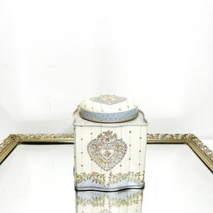 May include: A decorative metal tin with a lid, featuring a heart-shaped floral design. The tin has a cream-colored background with blue accents and a light blue lid. The tin is rectangular with rounded edges and is sitting on a mirrored surface.