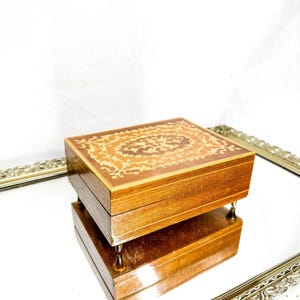 Peut inclure: Petite boîte à musique rectangulaire en bois, ornée d'un motif incrusté sur le couvercle. La boîte est de couleur marron chaud et repose sur quatre petits pieds argentés. Le couvercle présente un motif floral détaillé en tons de bois plus clairs.