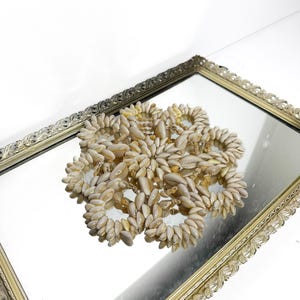 May include: A circular decorative mat constructed from numerous small, light beige cowrie shells arranged in a repeating, interconnected floral pattern. The shells are tightly clustered to form a series of linked rings, creating a textured, openwork design.