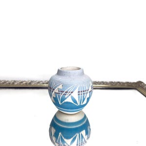 Puede incluir: Un pequeño jarrón redondo de cerámica azul y blanca con un patrón decorativo de hojas y líneas. El jarrón se refleja en una superficie espejada. El jarrón tiene una parte superior azul claro y una base azul más oscura.